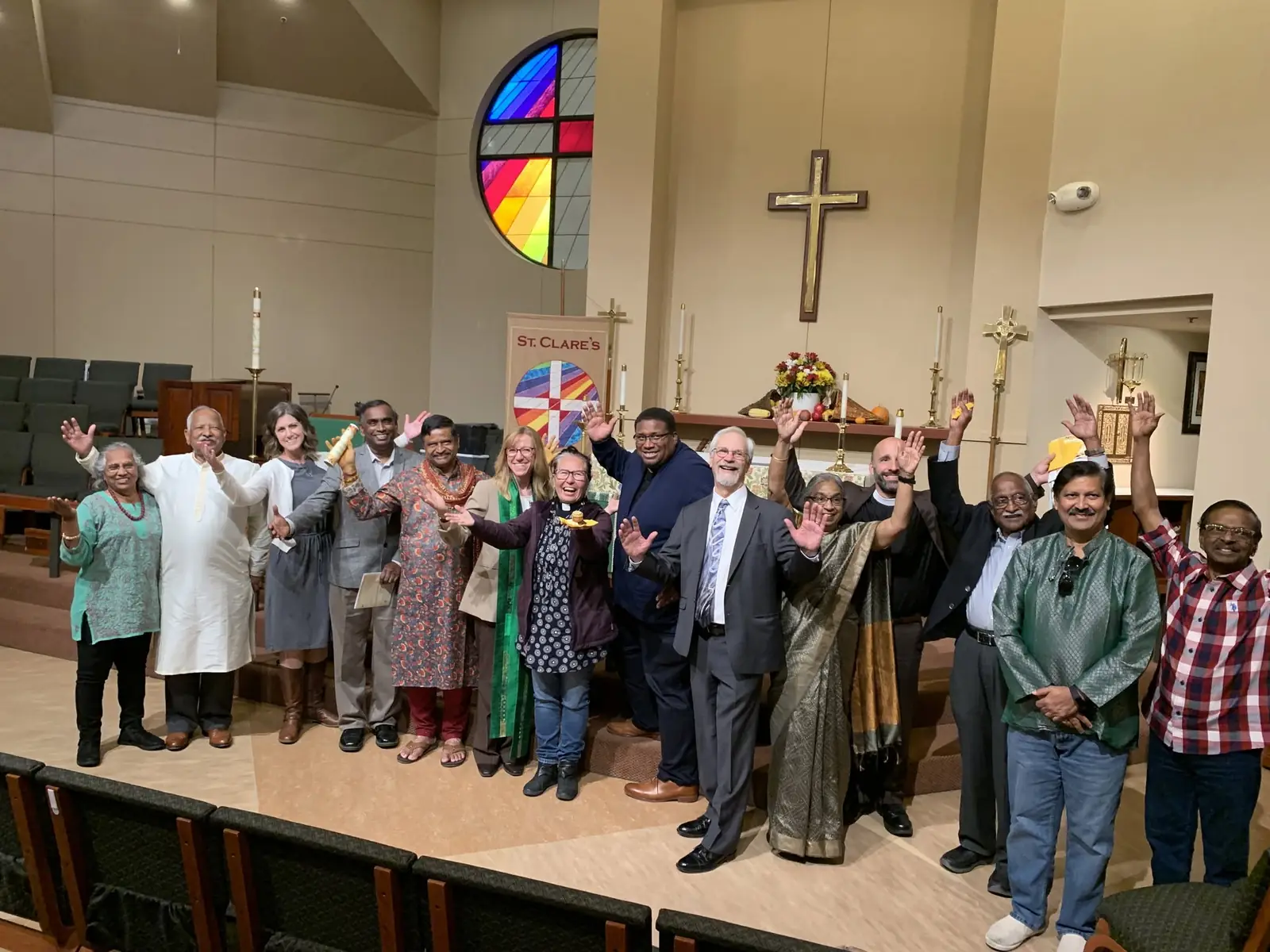 About fifteen people of different faiths and cultures, some in clergy vestments and some in saris and kurtas, smile and raise their hands together before the altar at St. Clare's.