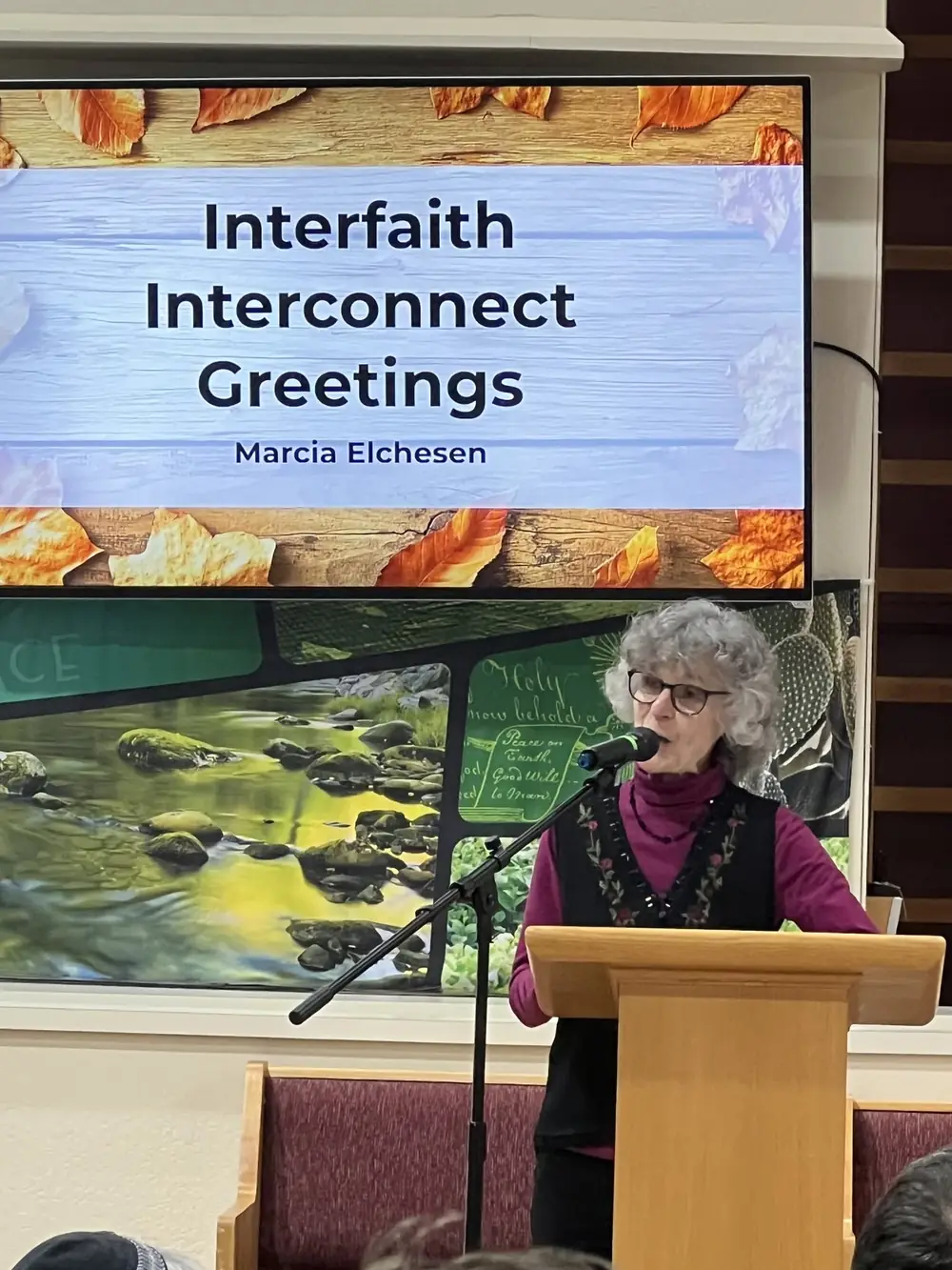 Marcia Elchesen speaks at a microphone beneath a screen reading Interfaith Interconnect Greetings.