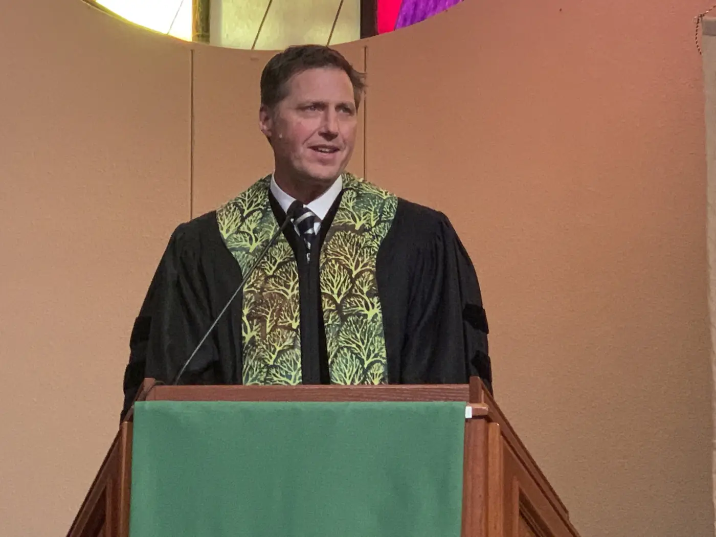 A speaker in a black robe and green stole addresses the room from a wooden pulpit.